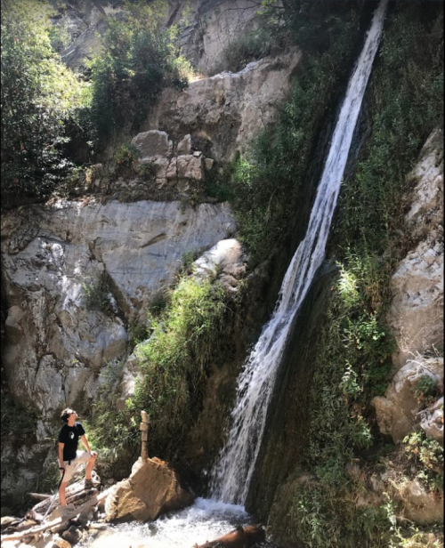 A person stands near a waterfall surrounded by rocky terrain and lush greenery.