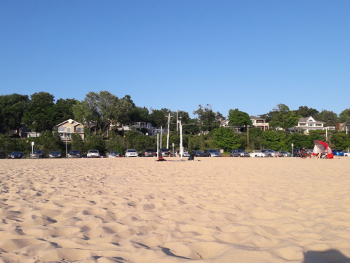 A sandy beach with parked cars in the background and a few beach setups under a clear blue sky.