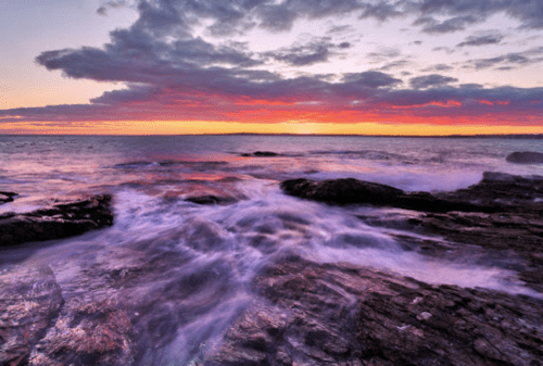 A serene seascape at sunset, with waves gently crashing over rocky shorelines under a colorful sky.