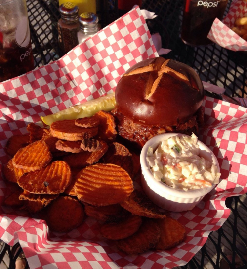 A plate with a pretzel bun burger, pickle, sweet potato chips, and a side of coleslaw in a white bowl.