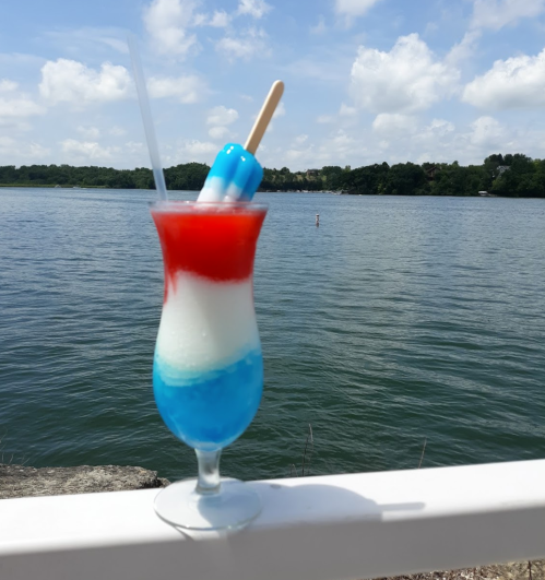 A colorful layered drink with red, white, and blue, topped with a popsicle, by a lake under a blue sky.