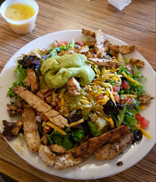 A colorful salad topped with grilled chicken, cheese, guacamole, and fresh vegetables on a white plate.