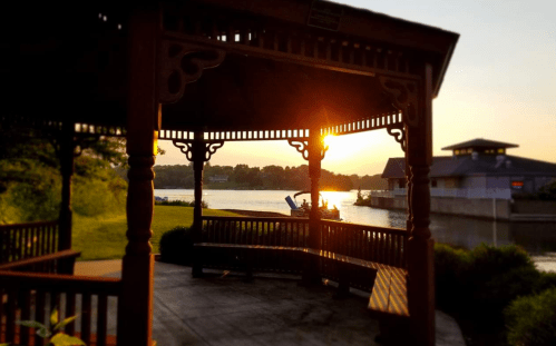 A gazebo by a river at sunset, with a silhouette of a person in a boat and buildings in the background.