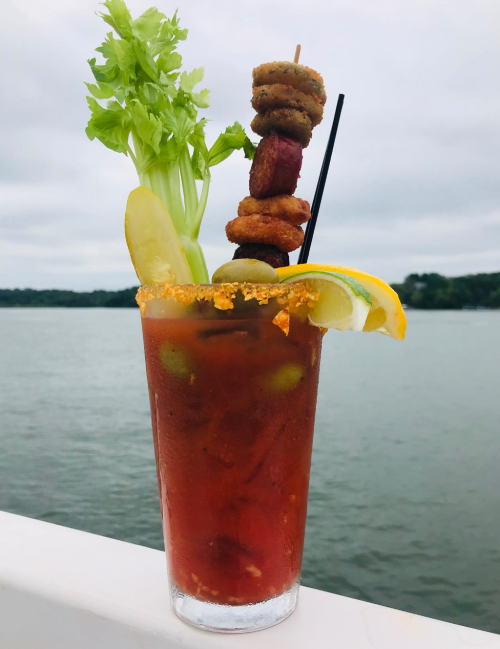 A vibrant Bloody Mary garnished with celery, pickles, olives, and skewered snacks, set against a water backdrop.