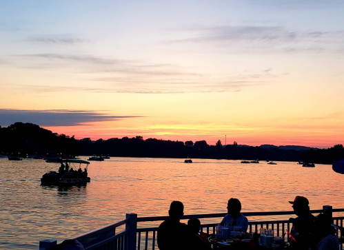 Silhouetted figures by a lake at sunset, with boats on the water and colorful sky reflecting on the surface.