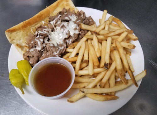 A plate with a sandwich filled with beef, drizzled with sauce, served with fries and a side of dipping sauce and peppers.