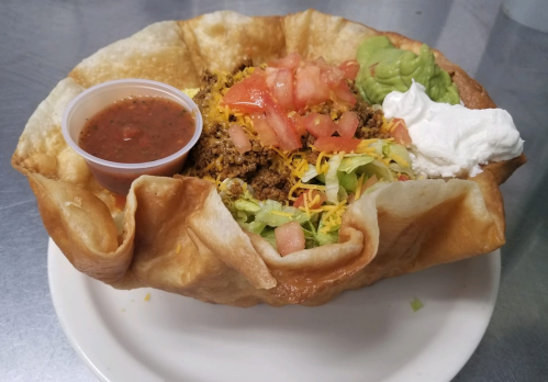 A taco salad in a crispy shell, filled with lettuce, ground beef, cheese, tomatoes, guacamole, sour cream, and salsa.