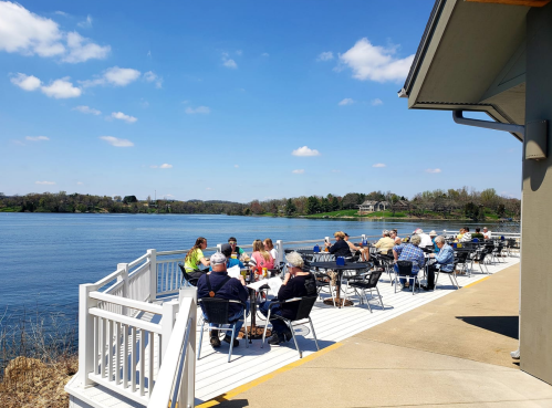 A group of people enjoying a sunny day on a deck by the water, with tables and chairs overlooking the lake.