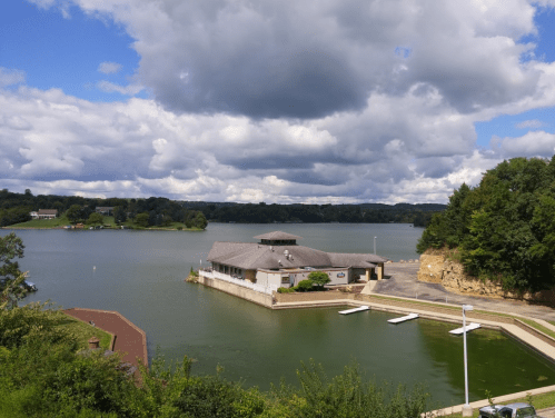 A serene lake view with a building on the shore, surrounded by green trees and cloudy skies.