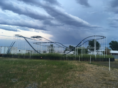 A roller coaster structure stands against a cloudy sky, surrounded by grass and trees.