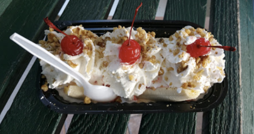 A banana split topped with whipped cream, chopped nuts, and cherries, served on a black tray with a white spoon.