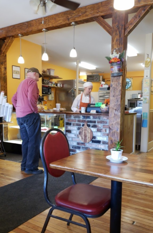 A cozy café interior with a customer at the counter and a barista in an apron, wooden beams, and a small table.