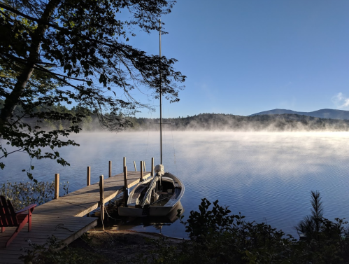 A serene lake at dawn with mist rising, a wooden dock, and a sailboat moored nearby. Trees frame the scene.