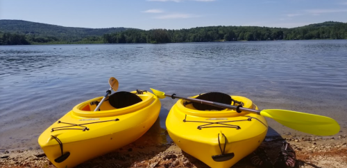 Two yellow kayaks on a sandy shore by a calm lake, surrounded by green hills under a clear blue sky.