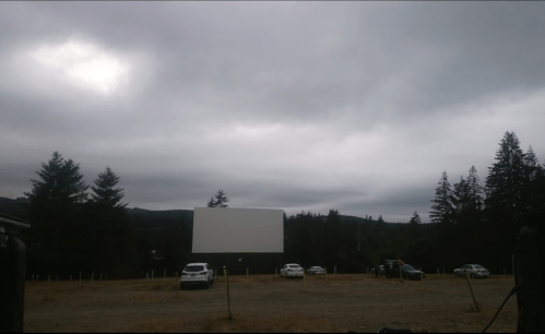 A cloudy sky over an empty outdoor movie screen with parked cars nearby and trees in the background.
