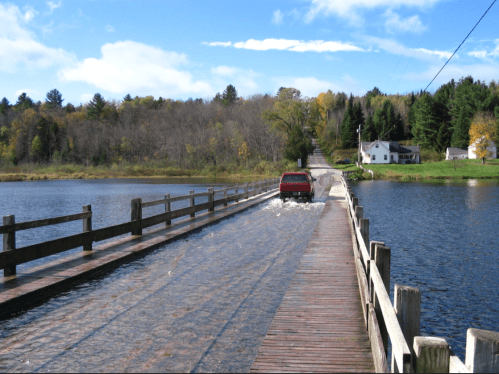 A red truck drives on a wooden bridge partially submerged in water, surrounded by trees and a house in the background.