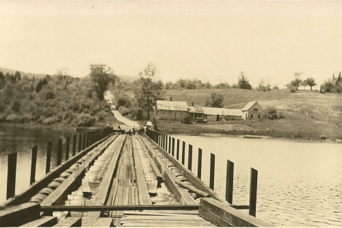 A vintage photo of a wooden bridge extending over a calm lake, leading to a rural landscape with a house in the distance.