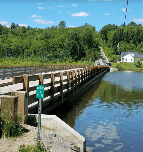 A wooden bridge spans a calm body of water, surrounded by lush greenery and a road leading into the distance.