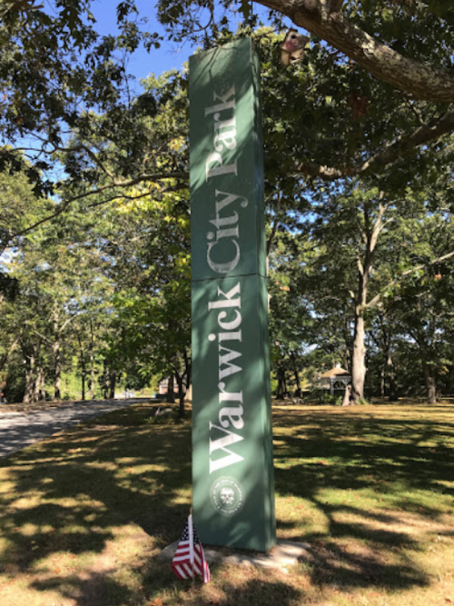 Sign for Warwick City Park surrounded by trees, with a small American flag at its base.