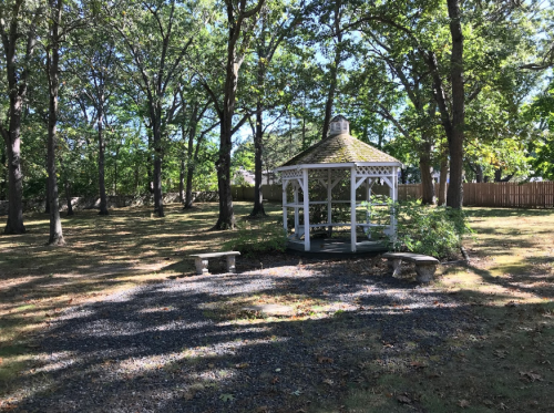 A white gazebo surrounded by trees in a grassy area, with stone benches nearby and dappled sunlight on the ground.