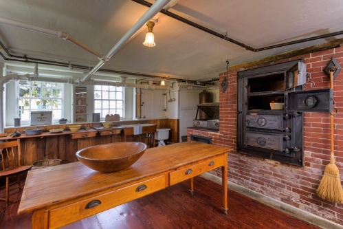 A rustic kitchen featuring a wooden table, brick walls, an antique stove, and large windows for natural light.