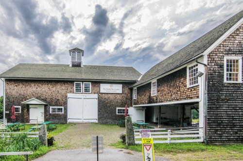 A rustic barn with a sign, surrounded by green grass and picnic tables, under a cloudy sky.