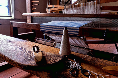 A wooden weaving loom with blue and white fabric, a spool of thread, scissors, and a shuttle on a rustic table.
