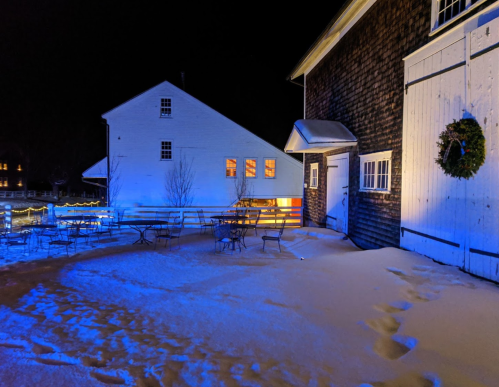 A snowy outdoor scene at night, featuring a barn, tables, and blue lighting, creating a serene winter atmosphere.
