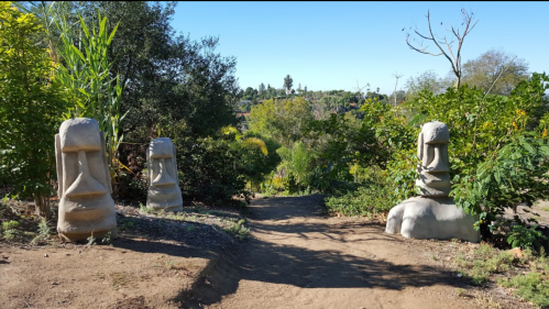 Two large stone statues resembling Easter Island moai stand along a dirt path surrounded by greenery.