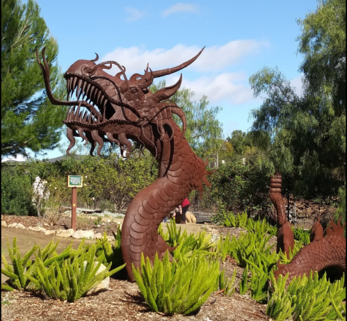 A large, rusted metal dragon sculpture coils among green plants under a blue sky.