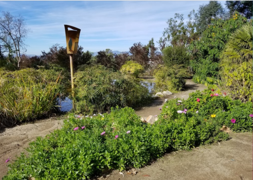 A serene garden scene with lush greenery, colorful flowers, and a wooden sculpture in the background under a clear sky.