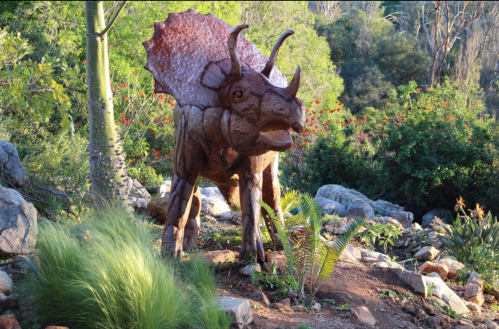 A lifelike sculpture of a Triceratops in a lush garden setting, surrounded by greenery and rocks.