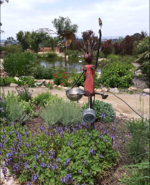 A whimsical garden sculpture made from kitchen utensils stands among vibrant flowers and greenery near a pond.