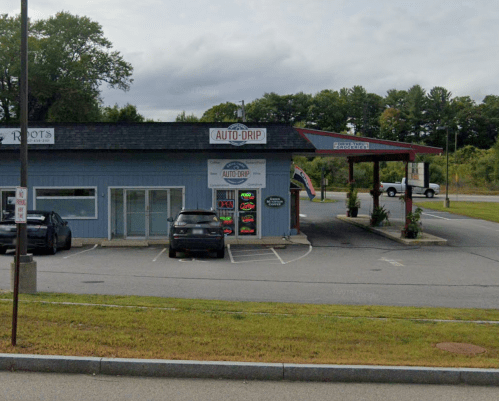 A small auto service building with a sign reading "Auto-Drip" and a gas station canopy nearby, surrounded by greenery.