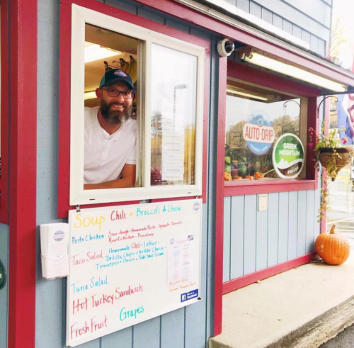 A man with a beard smiles from a food window, showcasing a menu of soups and sandwiches on a colorful sign.