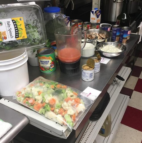 A kitchen countertop with various ingredients: vegetables, canned goods, a blender, and a bowl of cream.