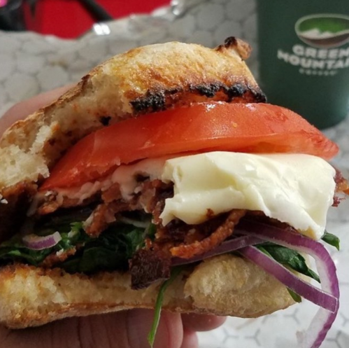 A close-up of a sandwich with layers of lettuce, tomato, onion, and meat on toasted bread, held in a hand.