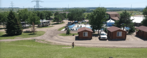 A scenic view of a campground with cabins, a pool, and trees, under a clear blue sky. A child is playing on the grass.