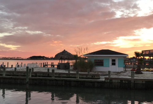 A serene waterfront scene at sunset, featuring a beach hut, palm trees, and a colorful sky reflected in the water.
