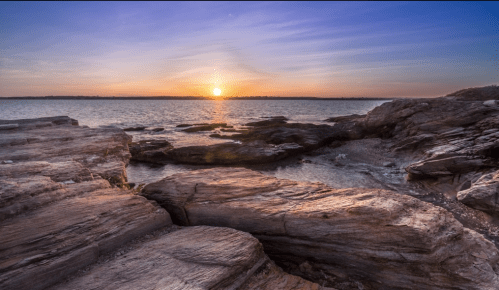 Sunset over rocky shoreline with calm water, creating a serene coastal landscape.