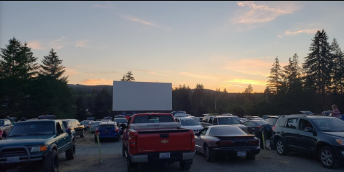 A drive-in movie theater at sunset, with cars parked in front of a large blank screen and trees in the background.