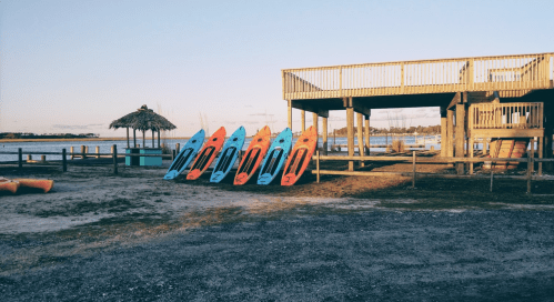 Colorful kayaks lined up on a sandy shore near a wooden deck and a thatched roof structure by the water.