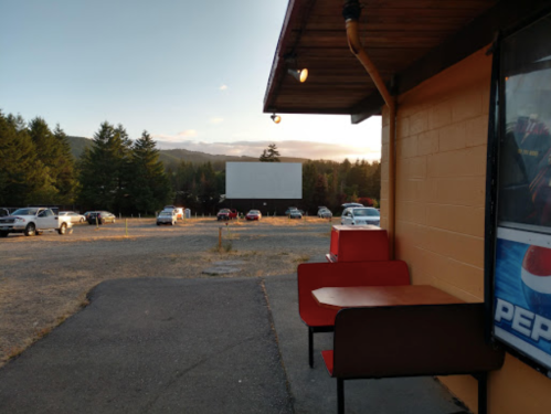 A view of a drive-in movie theater with parked cars and a large blank screen at sunset.