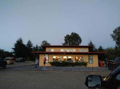 A two-story yellow building with a red roof, surrounded by trees, at dusk with people walking nearby.