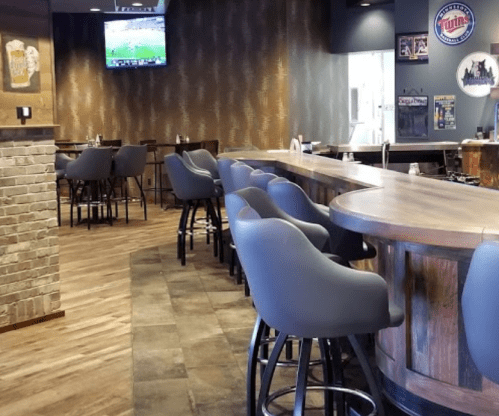 Modern bar interior with high stools, a wooden counter, and a TV showing a sports game. Warm lighting and decor.