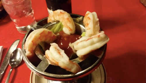 A metal dish with shrimp arranged around a bowl of cocktail sauce on a red tablecloth.