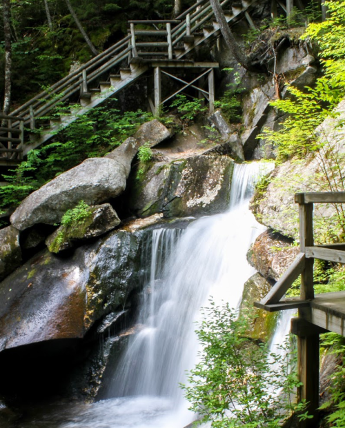 A serene waterfall cascades over rocks, surrounded by lush greenery and wooden stairs leading up the hillside.