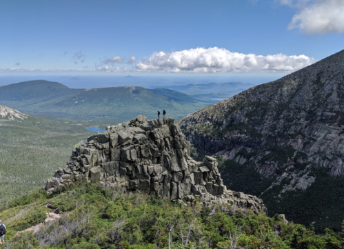 Two hikers stand atop a rocky outcrop, overlooking a lush valley and distant mountains under a partly cloudy sky.