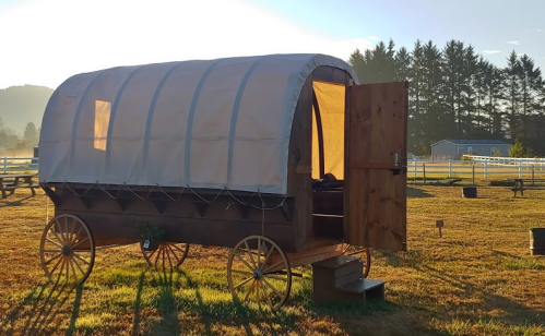 A wooden wagon with a canvas cover, parked in a grassy field at sunrise, surrounded by trees and a distant building.