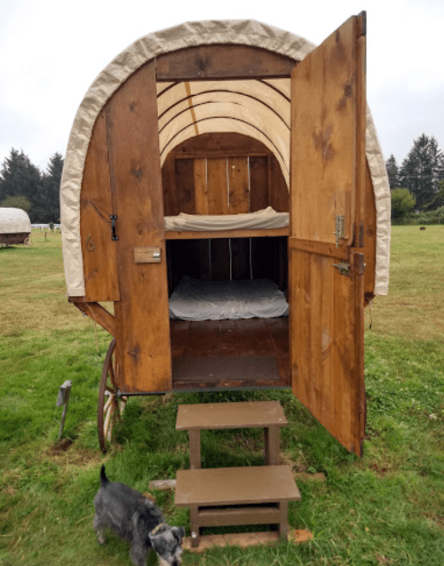 A wooden wagon with an arched roof, open doors revealing a simple interior with a bed, and steps leading up to it.